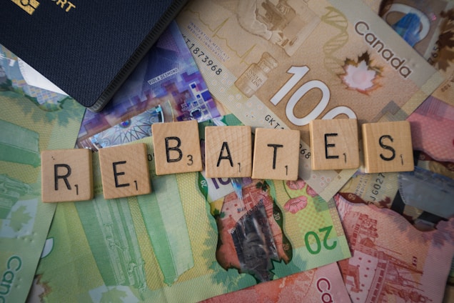 Scrabble tiles spelling out 'rebates' are arranged on top of various Canadian banknotes including denominations like 20 and 100. A part of a passport is visible in the corner.