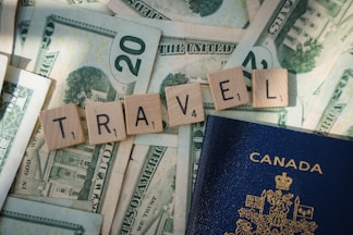 Wooden Scrabble tiles spelling 'TRAVEL' are arranged on top of US dollar bills, with a Canadian passport partially visible. The tiles and money suggest themes related to travel and finance.