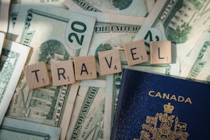 Wooden Scrabble tiles spelling 'TRAVEL' are arranged on top of US dollar bills, with a Canadian passport partially visible. The tiles and money suggest themes related to travel and finance.