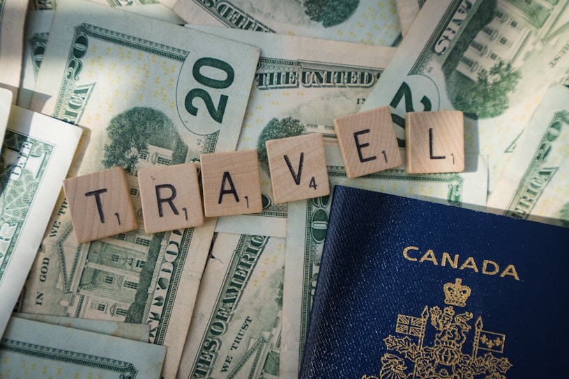 Wooden Scrabble tiles spelling 'TRAVEL' are arranged on top of US dollar bills, with a Canadian passport partially visible. The tiles and money suggest themes related to travel and finance.