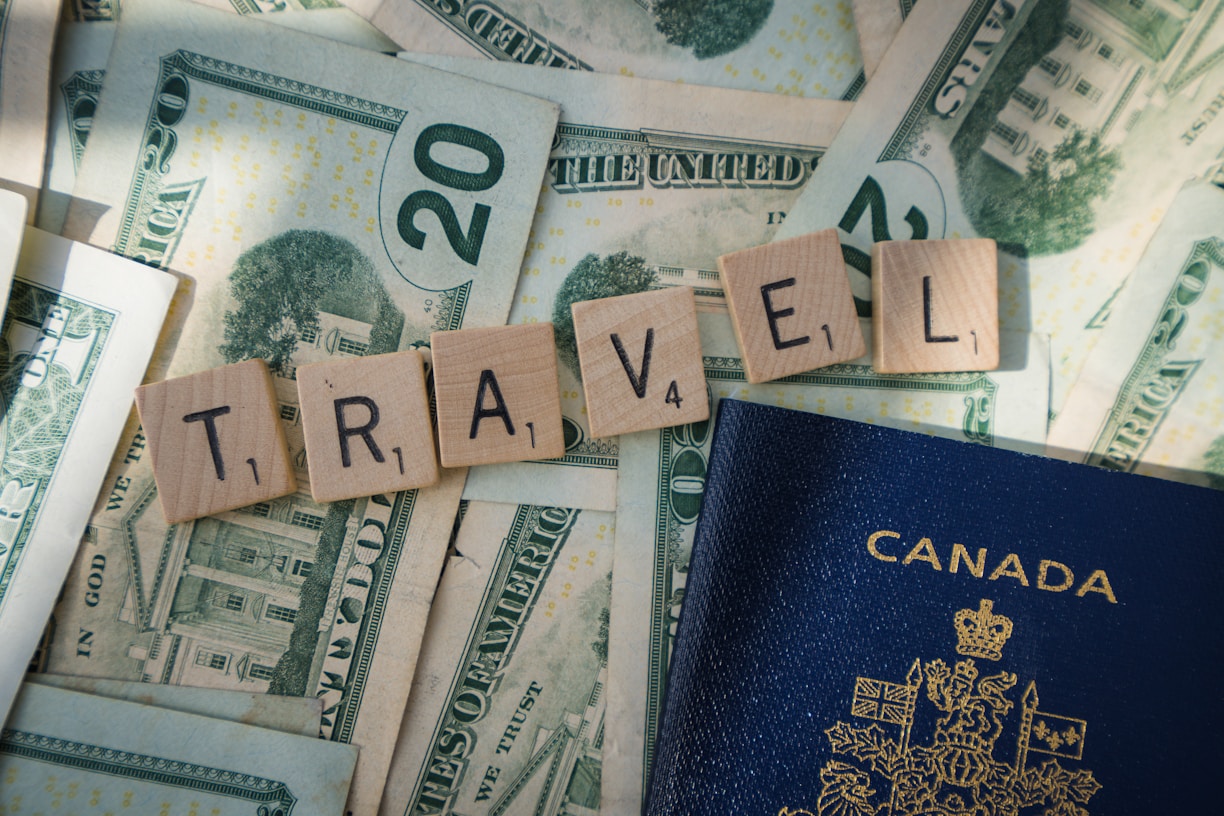 Wooden Scrabble tiles spelling 'TRAVEL' are arranged on top of US dollar bills, with a Canadian passport partially visible. The tiles and money suggest themes related to travel and finance.