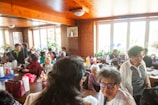 A group of people socializing in a warm, wood-paneled room with large windows letting in natural light. The room is filled with plants and framed photographs. People of varying ages are gathered around tables with food and drinks, engaged in conversation.