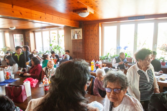 A group of people socializing in a warm, wood-paneled room with large windows letting in natural light. The room is filled with plants and framed photographs. People of varying ages are gathered around tables with food and drinks, engaged in conversation.