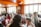 A group of people socializing in a warm, wood-paneled room with large windows letting in natural light. The room is filled with plants and framed photographs. People of varying ages are gathered around tables with food and drinks, engaged in conversation.