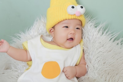 A baby wearing a knitted yellow hat with comical eyes and a small pink nose. The baby is dressed in a white garment with a yellow circle on it, resting on a fluffy white surface. The background is a soft green color.
