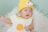 A baby wearing a knitted yellow hat with comical eyes and a small pink nose. The baby is dressed in a white garment with a yellow circle on it, resting on a fluffy white surface. The background is a soft green color.