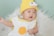 A baby wearing a knitted yellow hat with comical eyes and a small pink nose. The baby is dressed in a white garment with a yellow circle on it, resting on a fluffy white surface. The background is a soft green color.