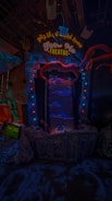 An illuminated theater entrance with a whimsical design, featuring a large open mouth above a sign that reads 'FEED ME THEATRE.' Bright, colorful lights surround the entrance, and various sea-themed decorations, including fish and tentacles, are present. The scene is dark with vibrant neon highlights, creating a playful and surreal atmosphere.