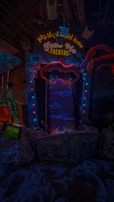 An illuminated theater entrance with a whimsical design, featuring a large open mouth above a sign that reads 'FEED ME THEATRE.' Bright, colorful lights surround the entrance, and various sea-themed decorations, including fish and tentacles, are present. The scene is dark with vibrant neon highlights, creating a playful and surreal atmosphere.