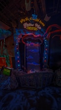 An illuminated theater entrance with a whimsical design, featuring a large open mouth above a sign that reads 'FEED ME THEATRE.' Bright, colorful lights surround the entrance, and various sea-themed decorations, including fish and tentacles, are present. The scene is dark with vibrant neon highlights, creating a playful and surreal atmosphere.