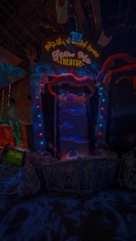 An illuminated theater entrance with a whimsical design, featuring a large open mouth above a sign that reads 'FEED ME THEATRE.' Bright, colorful lights surround the entrance, and various sea-themed decorations, including fish and tentacles, are present. The scene is dark with vibrant neon highlights, creating a playful and surreal atmosphere.