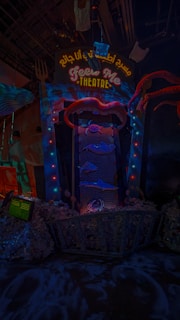 An illuminated theater entrance with a whimsical design, featuring a large open mouth above a sign that reads 'FEED ME THEATRE.' Bright, colorful lights surround the entrance, and various sea-themed decorations, including fish and tentacles, are present. The scene is dark with vibrant neon highlights, creating a playful and surreal atmosphere.