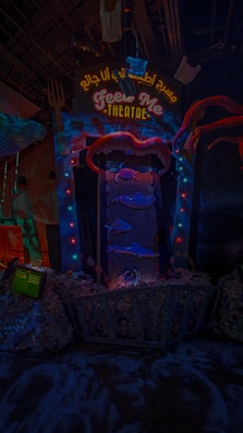 An illuminated theater entrance with a whimsical design, featuring a large open mouth above a sign that reads 'FEED ME THEATRE.' Bright, colorful lights surround the entrance, and various sea-themed decorations, including fish and tentacles, are present. The scene is dark with vibrant neon highlights, creating a playful and surreal atmosphere.