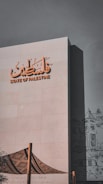 The image features a modern building facade with the words 'State of Palestine' in both Arabic and English. The structure is adorned with an artistic graphic, possibly depicting traditional architecture or cultural elements, in a subdued black-and-white style. In the foreground, there is a fabric structure featuring a traditional patterned design, supported by wooden poles.