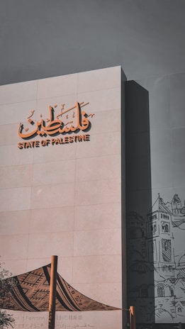The image features a modern building facade with the words 'State of Palestine' in both Arabic and English. The structure is adorned with an artistic graphic, possibly depicting traditional architecture or cultural elements, in a subdued black-and-white style. In the foreground, there is a fabric structure featuring a traditional patterned design, supported by wooden poles.