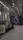 An indoor clothing store features a minimalist design with racks neatly arranged against a concrete floor. A variety of shirts and pants are displayed, primarily in neutral colors. On the right side, a large digital screen shows abstract artwork with vibrant purple and pink colors. The ceiling is adorned with exposed beams and track lighting, creating a modern industrial feel.