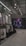 An indoor clothing store features a minimalist design with racks neatly arranged against a concrete floor. A variety of shirts and pants are displayed, primarily in neutral colors. On the right side, a large digital screen shows abstract artwork with vibrant purple and pink colors. The ceiling is adorned with exposed beams and track lighting, creating a modern industrial feel.