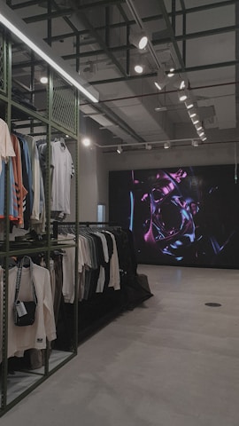 An indoor clothing store features a minimalist design with racks neatly arranged against a concrete floor. A variety of shirts and pants are displayed, primarily in neutral colors. On the right side, a large digital screen shows abstract artwork with vibrant purple and pink colors. The ceiling is adorned with exposed beams and track lighting, creating a modern industrial feel.