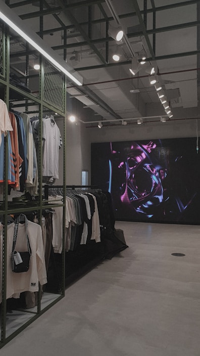 An indoor clothing store features a minimalist design with racks neatly arranged against a concrete floor. A variety of shirts and pants are displayed, primarily in neutral colors. On the right side, a large digital screen shows abstract artwork with vibrant purple and pink colors. The ceiling is adorned with exposed beams and track lighting, creating a modern industrial feel.