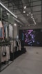 An indoor clothing store features a minimalist design with racks neatly arranged against a concrete floor. A variety of shirts and pants are displayed, primarily in neutral colors. On the right side, a large digital screen shows abstract artwork with vibrant purple and pink colors. The ceiling is adorned with exposed beams and track lighting, creating a modern industrial feel.