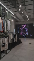 An indoor clothing store features a minimalist design with racks neatly arranged against a concrete floor. A variety of shirts and pants are displayed, primarily in neutral colors. On the right side, a large digital screen shows abstract artwork with vibrant purple and pink colors. The ceiling is adorned with exposed beams and track lighting, creating a modern industrial feel.