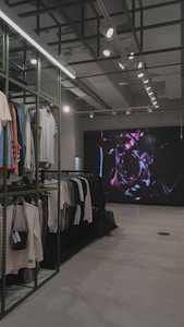 An indoor clothing store features a minimalist design with racks neatly arranged against a concrete floor. A variety of shirts and pants are displayed, primarily in neutral colors. On the right side, a large digital screen shows abstract artwork with vibrant purple and pink colors. The ceiling is adorned with exposed beams and track lighting, creating a modern industrial feel.