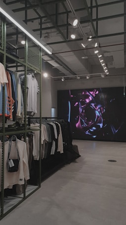 An indoor clothing store features a minimalist design with racks neatly arranged against a concrete floor. A variety of shirts and pants are displayed, primarily in neutral colors. On the right side, a large digital screen shows abstract artwork with vibrant purple and pink colors. The ceiling is adorned with exposed beams and track lighting, creating a modern industrial feel.