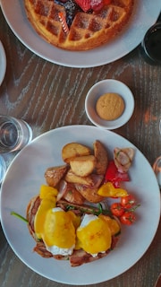A brunch spread featuring a plate with eggs Benedict topped with hollandaise, served alongside roasted potatoes, cherry tomatoes, and sliced peppers. Nearby is a large, round waffle adorned with fresh berries and a small candle. A glass of water, a small bowl with a cookie, and a cup of coffee are also present on the wooden table.