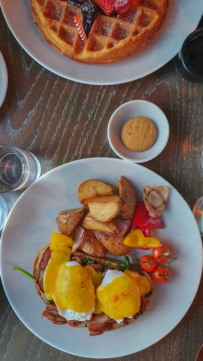 A brunch spread featuring a plate with eggs Benedict topped with hollandaise, served alongside roasted potatoes, cherry tomatoes, and sliced peppers. Nearby is a large, round waffle adorned with fresh berries and a small candle. A glass of water, a small bowl with a cookie, and a cup of coffee are also present on the wooden table.