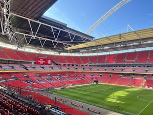 A large football stadium with red seating and an open roof structure. The seating area displays the word 'WEMBLEY' in large letters. A scoreboard shows 'Finalists 2022' with a team logo. The field is well-maintained with lush green grass, and a few people are visible walking on the pitch.