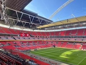 A large football stadium with red seating and an open roof structure. The seating area displays the word 'WEMBLEY' in large letters. A scoreboard shows 'Finalists 2022' with a team logo. The field is well-maintained with lush green grass, and a few people are visible walking on the pitch.