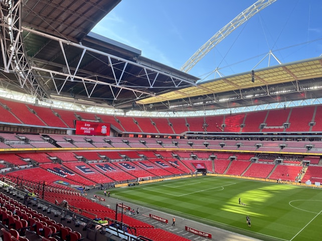 A large football stadium with red seating and an open roof structure. The seating area displays the word 'WEMBLEY' in large letters. A scoreboard shows 'Finalists 2022' with a team logo. The field is well-maintained with lush green grass, and a few people are visible walking on the pitch.