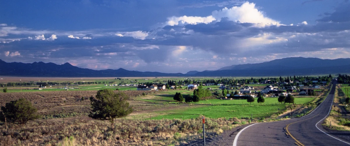 Green fields and houses outside Panguitch, Utah, beside a curving road, with mountains under storm clouds