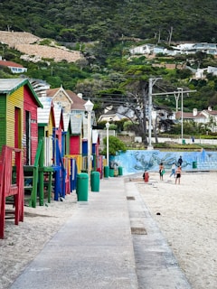 Colorful beach huts line the left side of a sandy path leading to the beach. The huts are painted in shades of red, blue, green, and yellow. In the background, green hills rise up with houses scattered on them. On the beach, a few people are gathered near a vivid mural on a wall.
