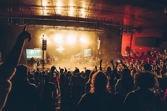 A lively concert scene with a large crowd facing the stage in a dimly lit auditorium. Bright stage lights illuminate the area, and people have their hands raised, possibly enjoying the music. There are musicians on stage and screens displaying visuals on both sides.
