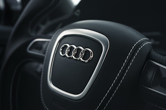 A close-up view of a car steering wheel featuring the Audi logo, with black leather and white stitching. The surroundings are out of focus, and the lighting accentuates the metallic elements.