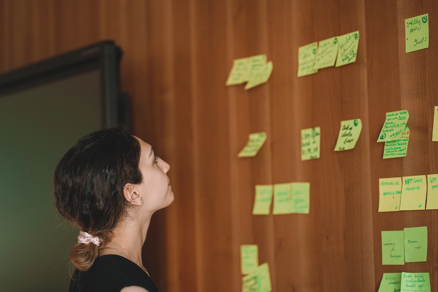 A woman with dark hair tied back standing close to a wooden wall of green notes, reading one of them