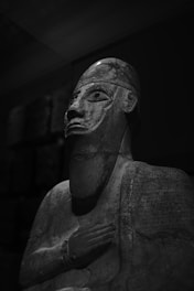 A grayscale image of an ancient statue depicting a human figure with a detailed, elongated face, wearing a headpiece. The statue's body is adorned with intricate carvings, possibly inscriptions or symbols. The lighting is dramatic, creating deep shadows and highlighting the detailed textures of the sculpture. The background is dark, which emphasizes the figure.