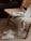 A cozy setting featuring a woven chair with a light gray blanket draped over it. On the chair rests a closed journal with a black pen on top. The chair is positioned on a textured, woven rug, and nearby are a pair of fluffy, cream-colored slippers.