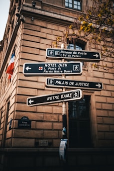 An old building with a formal architectural style serves as the backdrop for a cluster of street signs pointing towards various locations, including 'Bureau de Poste', 'Hotel Dieu', 'Palais de Justice', and 'Notre Dame'. Leaves from a nearby tree add a touch of nature to the urban setting. A small French flag is visible, adding a national touch.