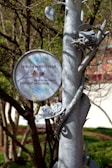 A metal pole with sculpted birds and leaves is adorned with a sign indicating park rules. The sign reads 'THIS IS A PASSIVE PARK' with instructions against skateboarding and littering. The scene is set against a backdrop of trees and greenery.
