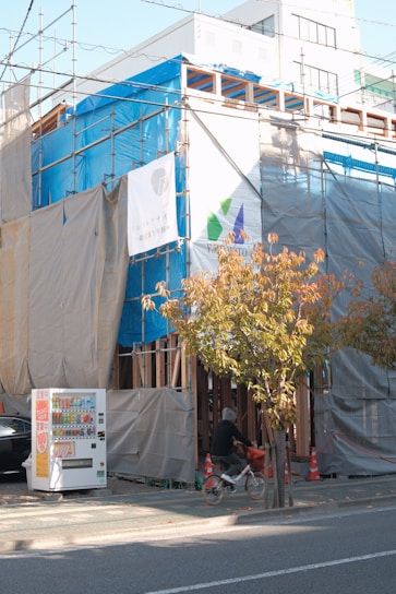 A building under construction is covered with blue and gray tarps. Scaffolding is visible, and two signs with logos and text are affixed to the coverings. In the foreground, a person rides a bicycle past a row of trees with autumn leaves. A vending machine is situated on the sidewalk next to a parked black car.