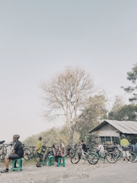 A group of cyclists are gathered at a resting stop by the side of the road. Several bicycles are parked nearby, and people wearing cycling gear are sitting on plastic stools. A leafless tree stands prominently in the background, and a small structure with a corrugated metal roof is visible. The scene appears relaxed and informal.