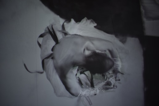 A black and white artistic image of a dancer elegantly captured in motion. The figure is curled inward, with flowing garments that suggest dynamic movement and grace. The focus is on the blurred, ethereal quality of the movement, providing a sense of fluidity.