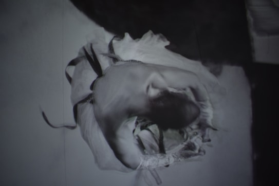 A black and white artistic image of a dancer elegantly captured in motion. The figure is curled inward, with flowing garments that suggest dynamic movement and grace. The focus is on the blurred, ethereal quality of the movement, providing a sense of fluidity.
