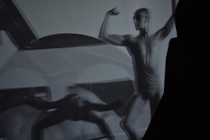 A monochrome image of a person in a dynamic pose, showcasing a muscular figure. The image has overlapping shadows, creating an abstract and artistic effect. The focus is on the strength and form of the human body, emphasizing contours and muscle definition.