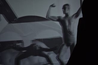 A monochrome image of a person in a dynamic pose, showcasing a muscular figure. The image has overlapping shadows, creating an abstract and artistic effect. The focus is on the strength and form of the human body, emphasizing contours and muscle definition.