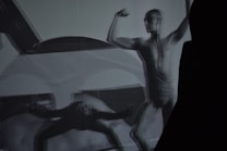 A monochrome image of a person in a dynamic pose, showcasing a muscular figure. The image has overlapping shadows, creating an abstract and artistic effect. The focus is on the strength and form of the human body, emphasizing contours and muscle definition.