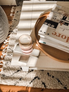 A cozy and stylish living space features a light wooden coffee table with a stack of magazines on a round tray, alongside a frothy drink in a pink glass on top of two coasters. The table sits on a textured, patterned rug that covers a wooden floor. Natural light casts gentle shadows across the table, adding warmth to the scene.