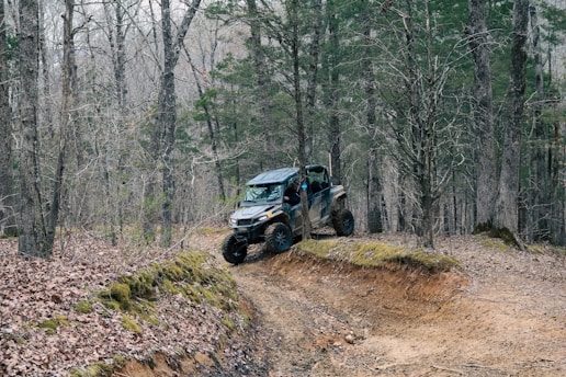 A rugged off-road vehicle is navigating a muddy trail through a dense forest. The scene is surrounded by tall trees with bare branches, indicating a winter or early spring setting. The ground is covered with fallen leaves and patches of moss.